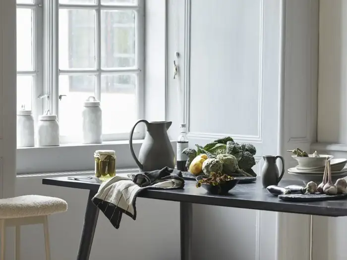 Et køkkenbord med grøntsager, tallerkener, krukker, en kande og et håndklæde nær et vindue med hvide dunke i vindueskarmen.