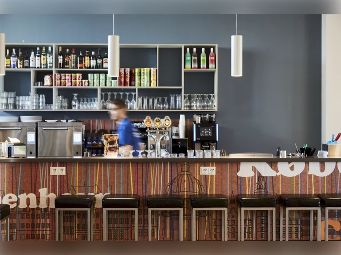 En moderne cafébar med hylder med drinks og glas bag disken, barstole foran og en person, der arbejder bag baren.