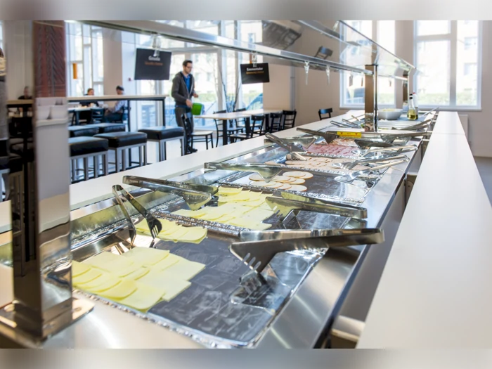 En selvbetjeningsbuffet med bakker med skiveskåret ost og kødpålæg; en person med en bakke går i baggrunden.