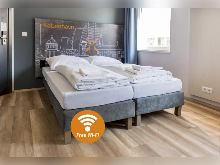 Et moderne hotelværelse med to enkeltsenge skubbet sammen, pænt opredt med hvidt sengetøj; på et skilt står der Free Wi-Fi, og på et vægmaleri står der København.