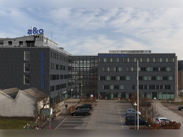 En moderne hotelbygning med a&o hostels-skiltning, store vinduer, parkerede biler foran og en delvis overskyet himmel over.