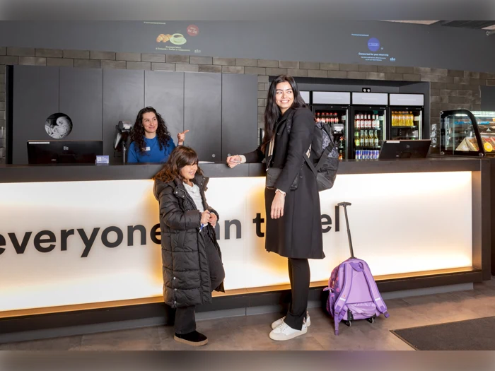 En kvinde og en pige med en lilla kuffert står ved en hotelreception og smiler til en receptionist bag skranken.