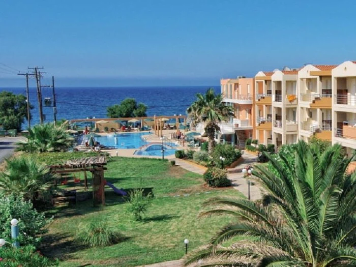 Et feriested ved havet med pool, liggestole, palmer og et stort græsareal med orange og beige bygninger med udsigt over det blå hav.