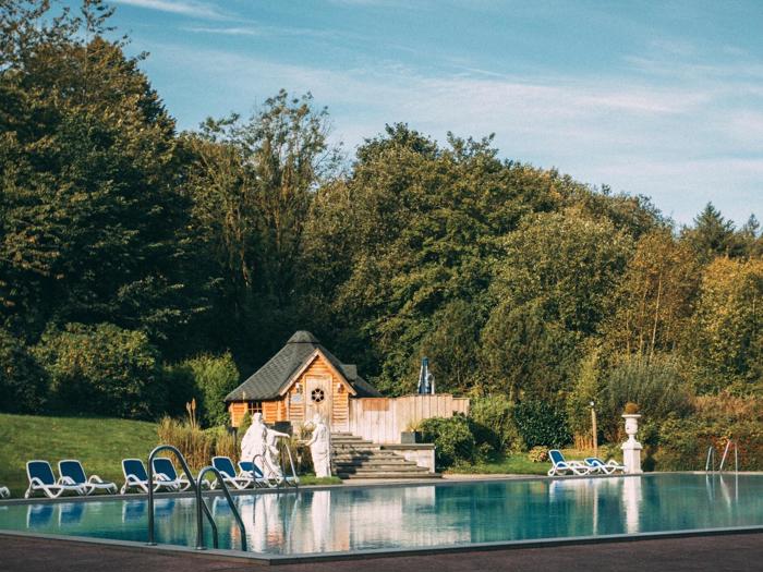 En udendørs swimmingpool med liggestole, statuer og en lille træhytte omgivet af frodige grønne træer under en klar blå himmel.