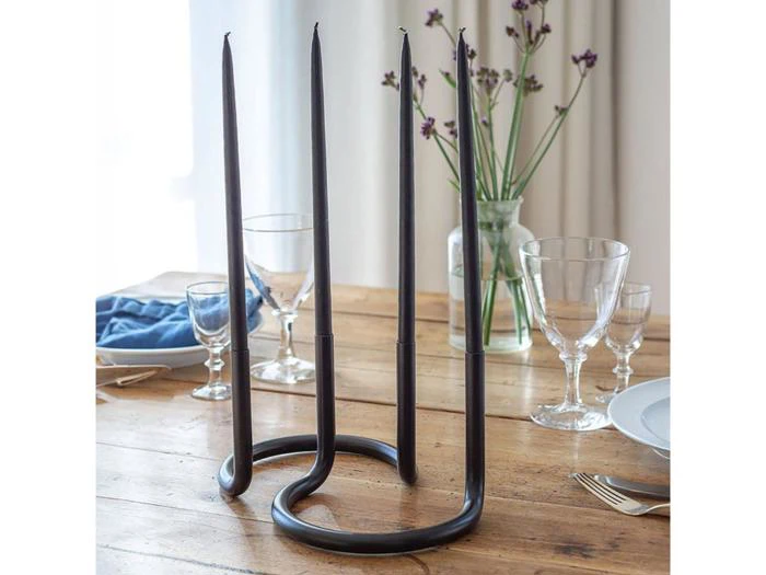 En moderne lysestage i sort metal med tre høje sorte lys på et spisebord i træ dækket med glas, tallerkener og en blå serviet; i baggrunden ses blomster og gardiner.