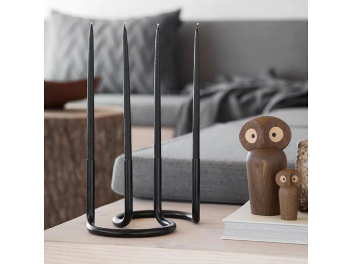 En moderne sort lysestage med fire høje stearinlys står på et bord ved siden af to uglefigurer i træ og en bog; en sofa og en træskammel står i baggrunden.