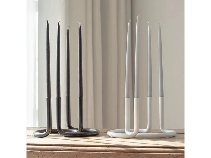 Fire U-formede lysestager, to sorte og to hvide, der hver især holder høje, slanke fyrfadslys, placeret på en træplade ved et vindue med gennemsigtige gardiner.