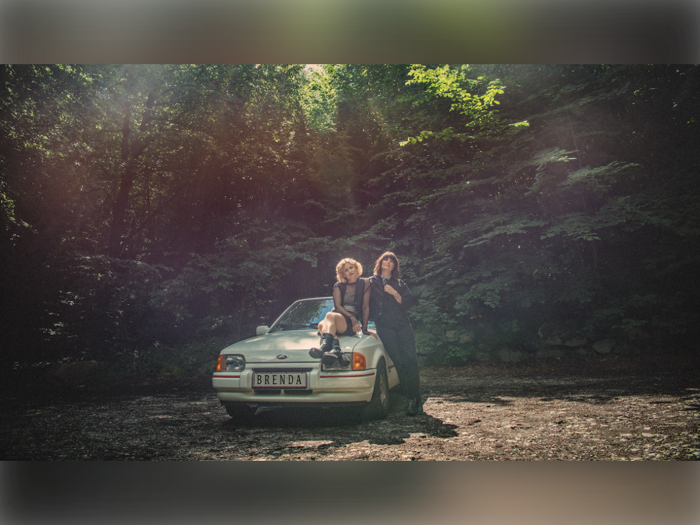 To personer poserer ved en bil i en solbeskinnet skov; den ene sidder på kølerhjelmen, den anden står ved siden af den. På bilens nummerplade står der BRENDA.