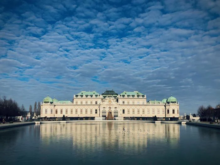 Et stort, udsmykket palads med grønt tag spejler sig i en stille dam under en himmel fyldt med spredte skyer.