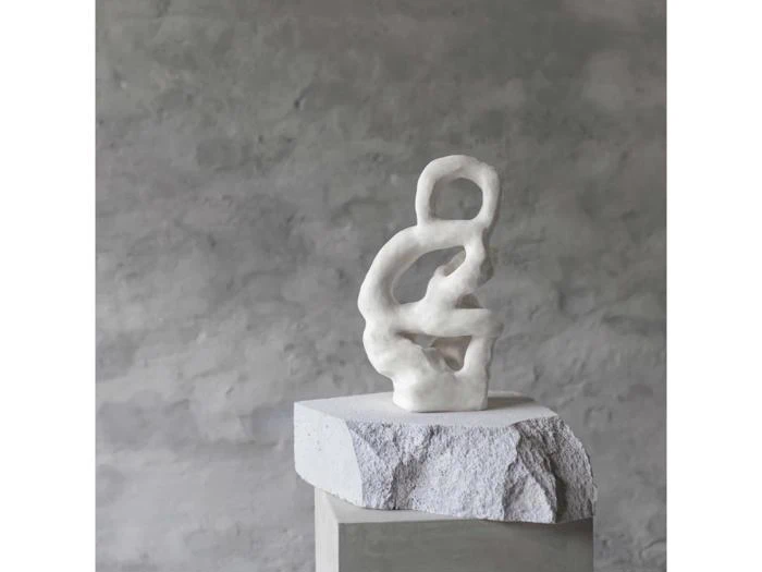Abstrakt hvid skulptur med slyngede, organiske former står på en grov stensokkel mod en grå, struktureret vægbaggrund.