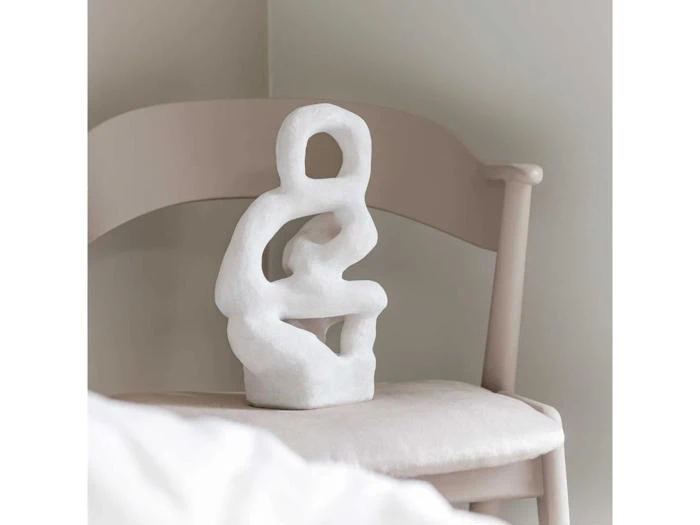 En hvid abstrakt skulptur med afrundede former hviler på en lys beige stol i et blødt oplyst, minimalistisk rum.