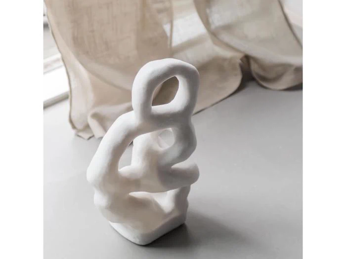 Hvid, abstrakt keramikskulptur med sammenflettede sløjfer står på en grå flade nær gennemsigtige, beige gardiner med blødt, naturligt lys fra et vindue i baggrunden.