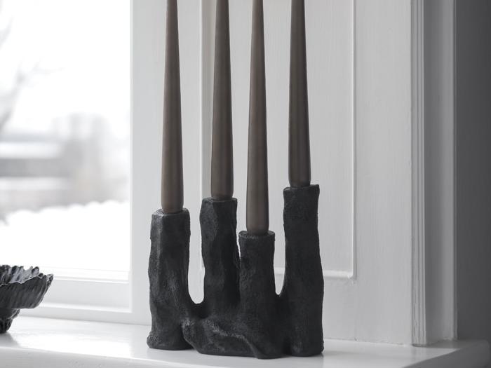 En sort, skulpturel lysestage med fire høje, mørke fyrfadslys står i en hvid vindueskarm foran et vindue.