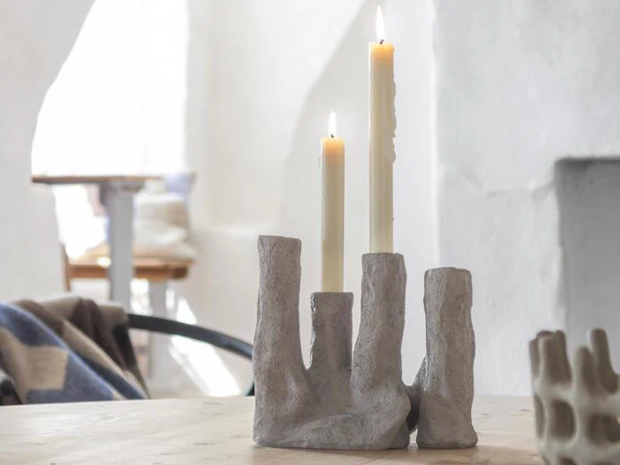 To tændte stearinlys i en abstrakt, grå stenlysestage står på et træbord i et svagt oplyst, minimalistisk rum.