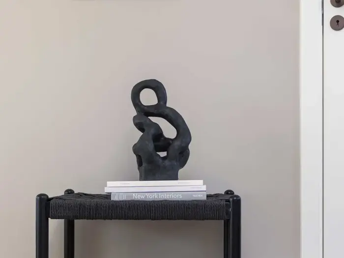 Abstrakt sort skulptur står på et lille bord med to stablede bøger mod en almindelig beige væg i en minimalistisk, moderne indretning.