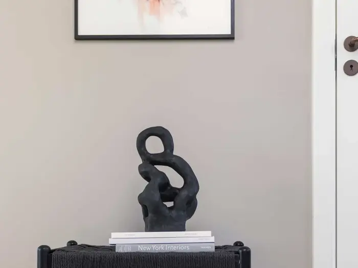 Abstrakt sort skulptur på et sort bord med stablede bøger, bl.a. New York Interiors, mod en lys beige væg og et delvist indrammet billede ovenover.