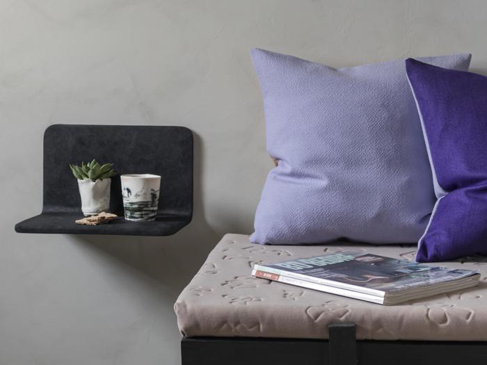 En hyggelig bænk med lilla og lilla puder, et magasin og en væghylde med en lille plante og to kopper mod en lysegrå væg.