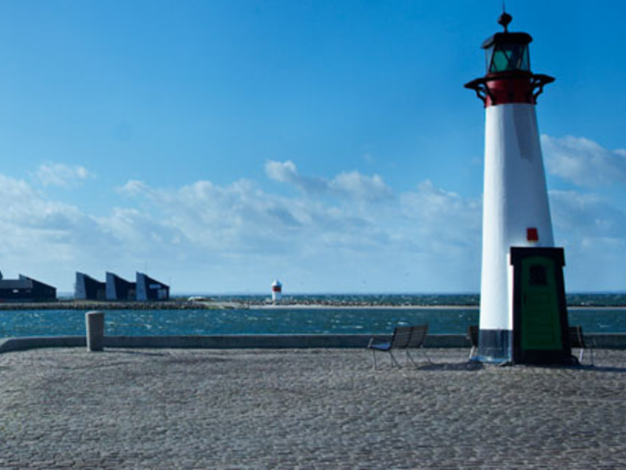 Et fyrtårn står på en brostensbelagt mole ud til havet, med bænke i nærheden og et andet fyrtårn, der kan ses i det fjerne under en blå himmel.