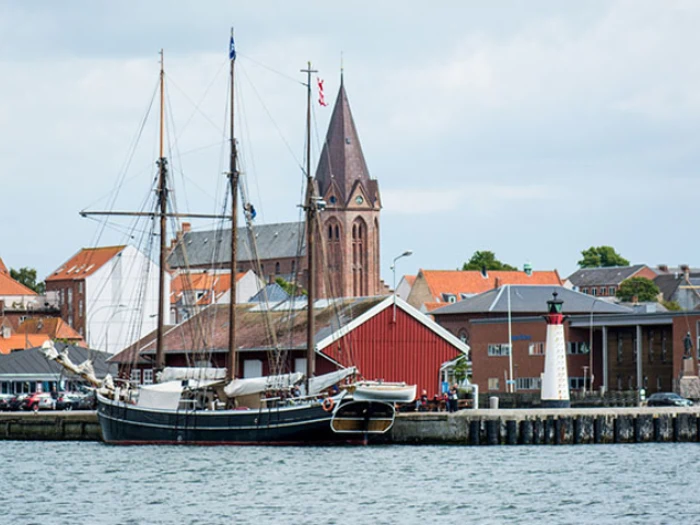 Et højt sejlskib ligger til kaj i en havn med bygninger med røde tage, en kirke med tårn og et hvidt fyrtårn i baggrunden.