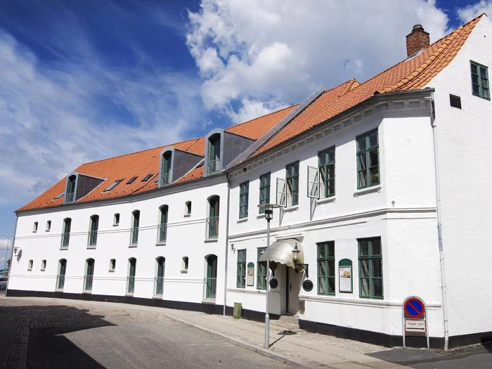 En hvid, toetagers bygning med grønne vinduer og et orange tegltag under en delvis overskyet blå himmel. Et skilt med parkeringsforbud er synligt foran.