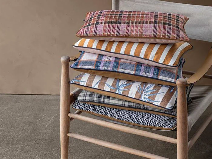 En træstol med fem farverige, mønstrede puder stablet på sædet, herunder ternede, stribede og geometriske mønstre.