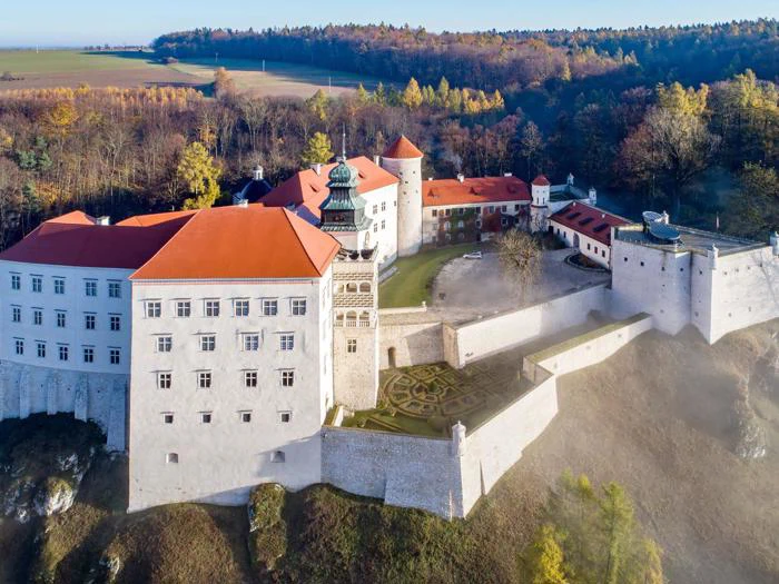 Luftfoto af et stort slot med røde tegltage, tårne og haver, omgivet af træer og landskab i efteråret.