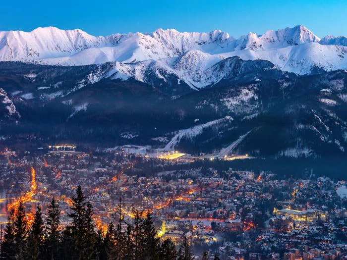 En by med lysende gadelygter beliggende i en dal, omgivet af snedækkede bjerge under en klar aftenhimmel.