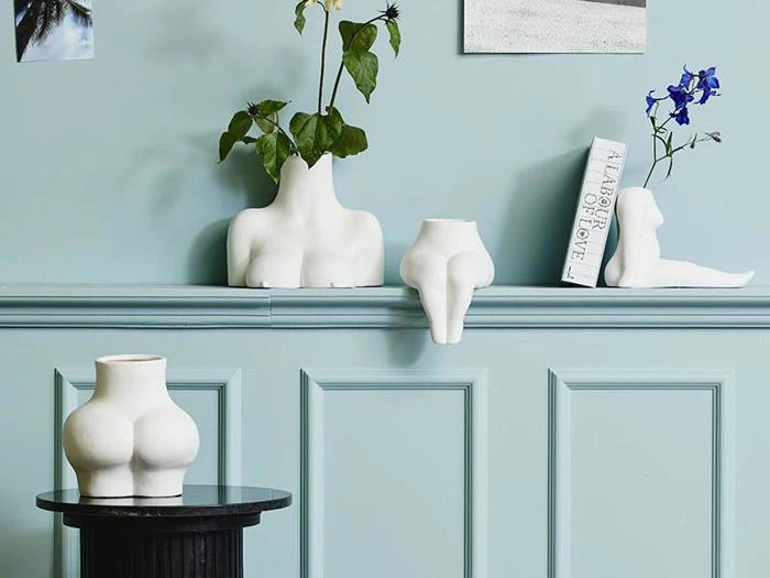 Minimalistiske hvide vaser formet som abstrakte menneskelige overkroppe og ben, udstillet på en lyseblå væg og et sort bord med blomster og en bog.