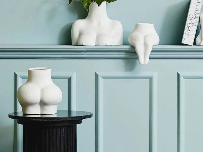 Tre hvide keramikvaser formet som abstrakte menneskekroppe er udstillet mod en lyseblå panelvæg med en vase på en sort piedestal og to på en afsats.