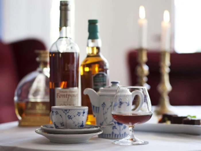 Et bord med spiritusflasker, en tekop, en tekande, et glas med brun væske og to tændte stearinlys i baggrunden.