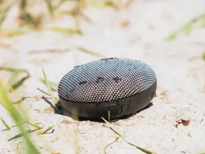 En lille bærbar Bluetooth-højttaler hviler på sandjord med nogle grønne græsstrå i nærheden.