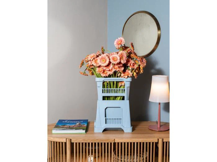 En buket lyserøde blomster i en lyseblå kasse står på et træbord ved siden af bøger, en lampe og et rundt spejl på en blå og hvid væg.