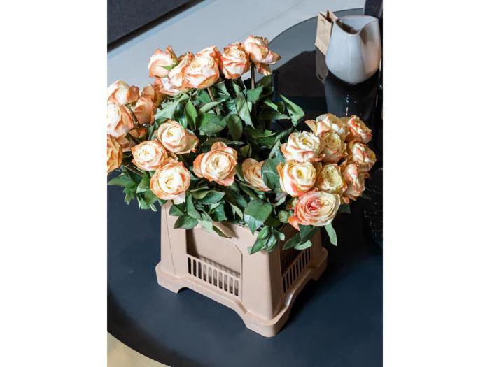 En buket ferskenfarvede og cremefarvede kunstige roser med grønne blade arrangeret i en beige plastkurv på et mørkt rundt bord.