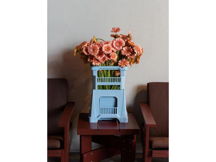 En buket lyserøde blomster i en blå plastikkasse står på et lille brunt bord mellem to brune lænestole op ad en lys væg.