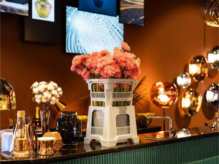 En buket lyserøde blomster i en hvid plastkasse står på en disk, omgivet af pyntegenstande og oplyst af varm, moderne belysning i et stilfuldt rum.