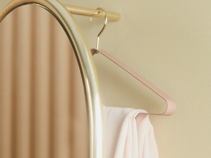 Et rundt spejl reflekterer en beige væg ved siden af en lyserød bøjle, der holder et hvidt stof på en træstang.