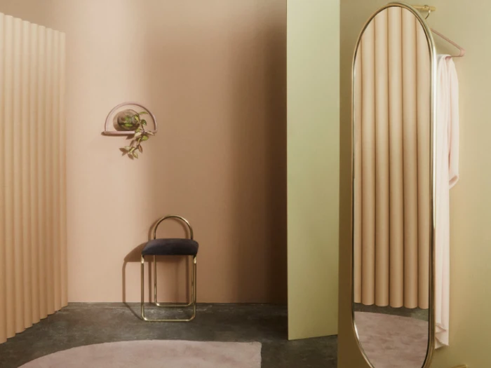 Minimalistisk rum med beige vægge, en lille skammel i sort og guld, en rund væghylde med en plante, et stort ovalt spejl og et blødt, naturligt lys.