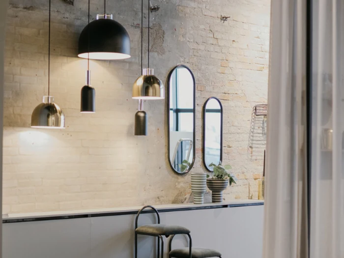 Moderne køkken med synlig murstensvæg, hængende pendler, tre ovale spejle, stablede skåle, en plante og en sort skammel ved køkkenbordet.