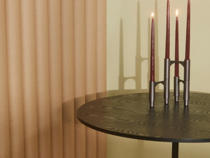 Et rundt bord i mørkt træ med en moderne lysestage med tre høje røde stearinlys, placeret ved siden af beige gardiner.