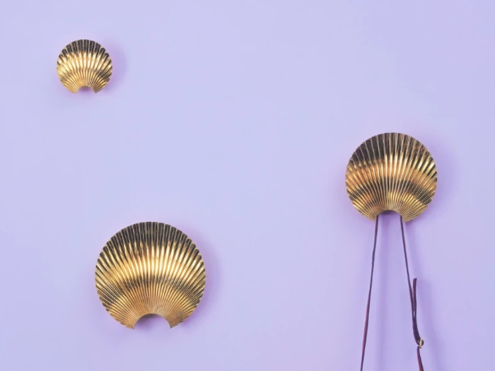 Tre gyldne, vifteformede vægdekorationer på en lyslilla baggrund. De to mindre er til venstre, og den største er til højre med to pinde hængende under sig.