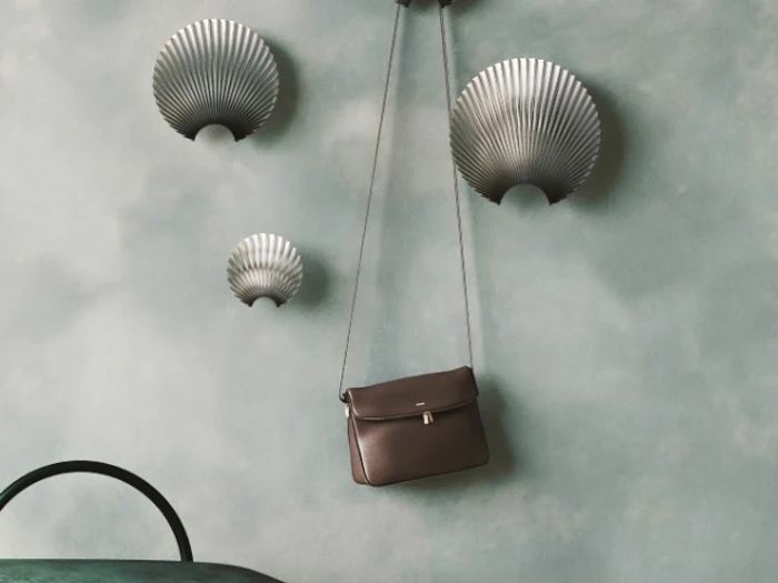En brun håndtaske hænger på en væg med tre dekorative, muslingeformede væglampetter i sølv mod en dæmpet blå baggrund.