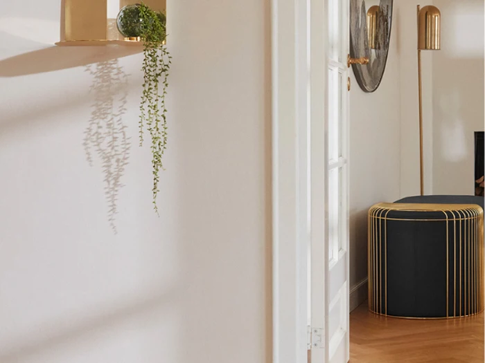 Minimalistisk indretning med en væghylde i guld med en lille plante, en sort ottoman med gulddetaljer og en gulvlampe i guld på et lyst trægulv.