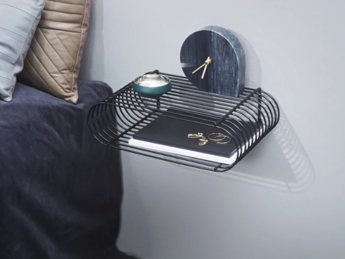 En moderne væghylde i sort tråd rummer en lille grøn skål, et mørkt marmorur, smykker og en lukket notesbog ved siden af en seng med grå og beige puder.