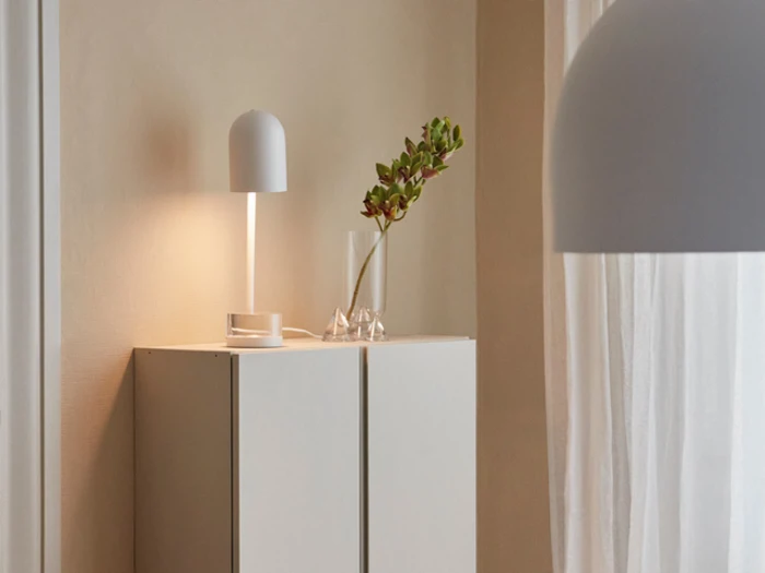 Minimalistisk rum med et hvidt skab, en moderne hvid bordlampe, en klar vase med en enkelt gren og gennemsigtige hvide gardiner, der lukker blødt lys ind.