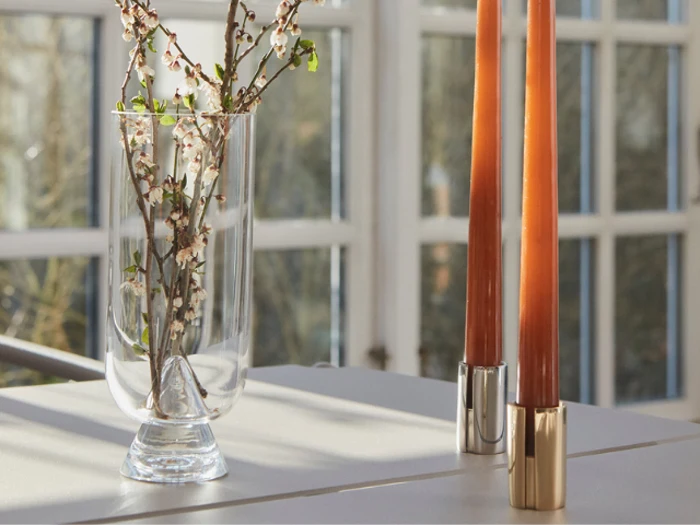 En klar glasvase med kirsebærblomstergrene og to høje orange stearinlys i metalholdere står på et solbeskinnet bord ved store vinduer.