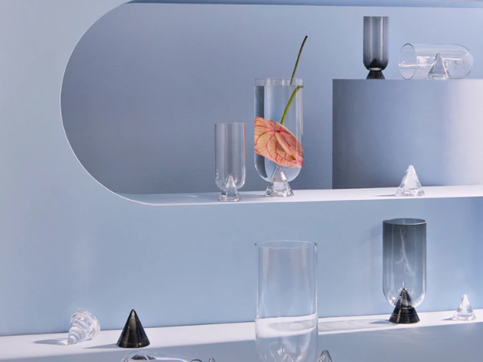 Minimalistisk udstilling af klare glasvaser og kegleformede genstande på forskudte hylder, hvor den ene vase indeholder en lyserød anthuriumblomst mod en lyseblå baggrund.