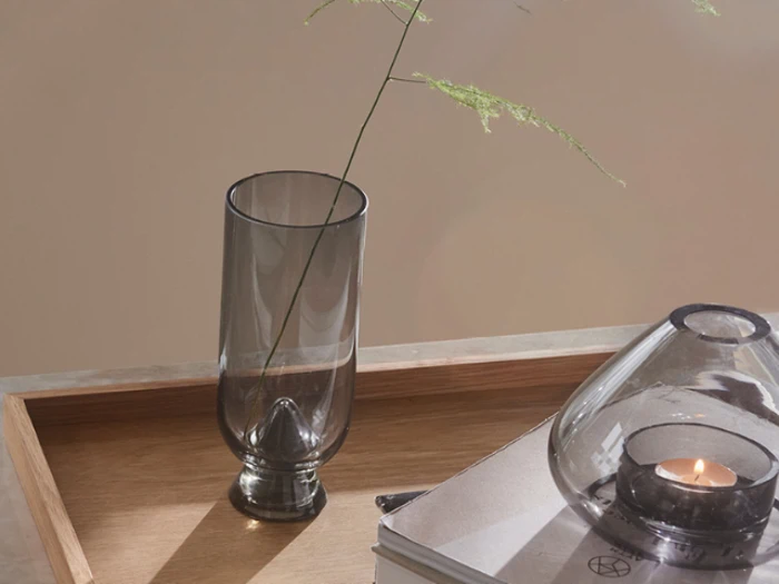 En klar glasvase med en enkelt tynd grøn stilk står på en træbakke ved siden af en lysestage og en lukket bog.