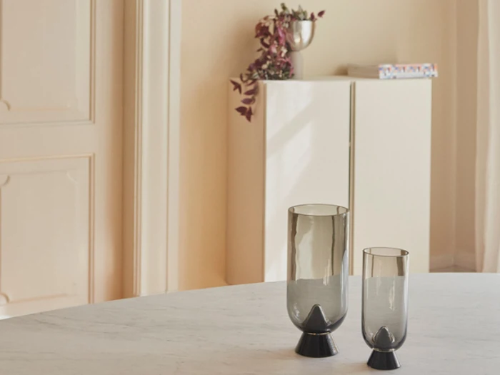 To moderne glasvaser med sort bund står på et rundt, hvidt marmorbord; i den slørede baggrund ses et hvidt skab med en sølvkop og en plante.