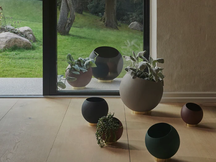 Moderne runde plantekasser med grønne planter står på et trægulv inde og ude ved en glasdør, hvor man kan se en græsklædt gård og træer udenfor.