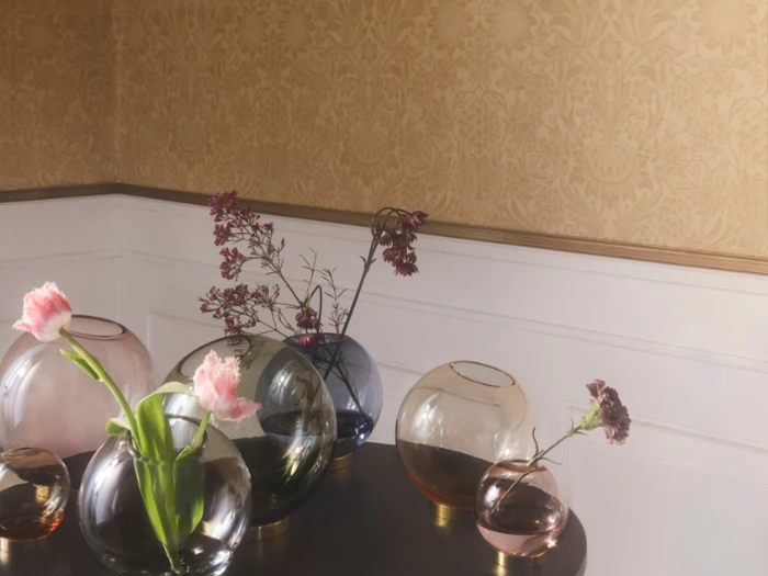 En gruppe runde glasvaser med lyserøde blomster og grønt står på et mørkt træbord mod en beige mønstret væg og hvid vægbeklædning.